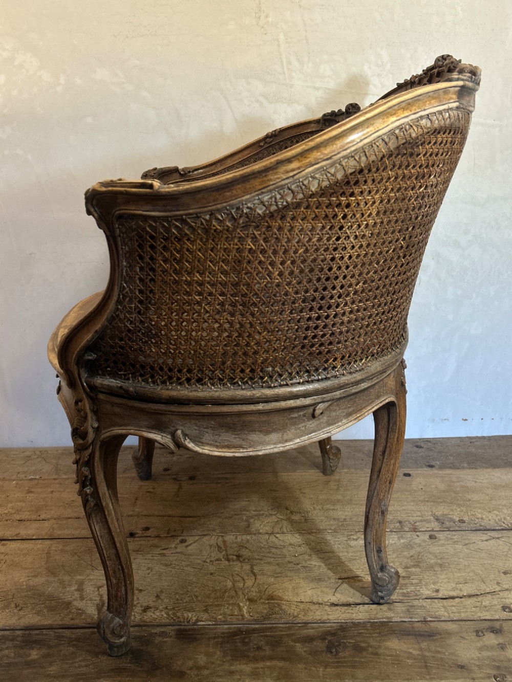 A 19th-century French Louis XV style Corbeille armchair featuring a curved hand-woven cane backrest, carved wooden frame with cabriole legs, and a beige upholstered seat cushion. View XIV