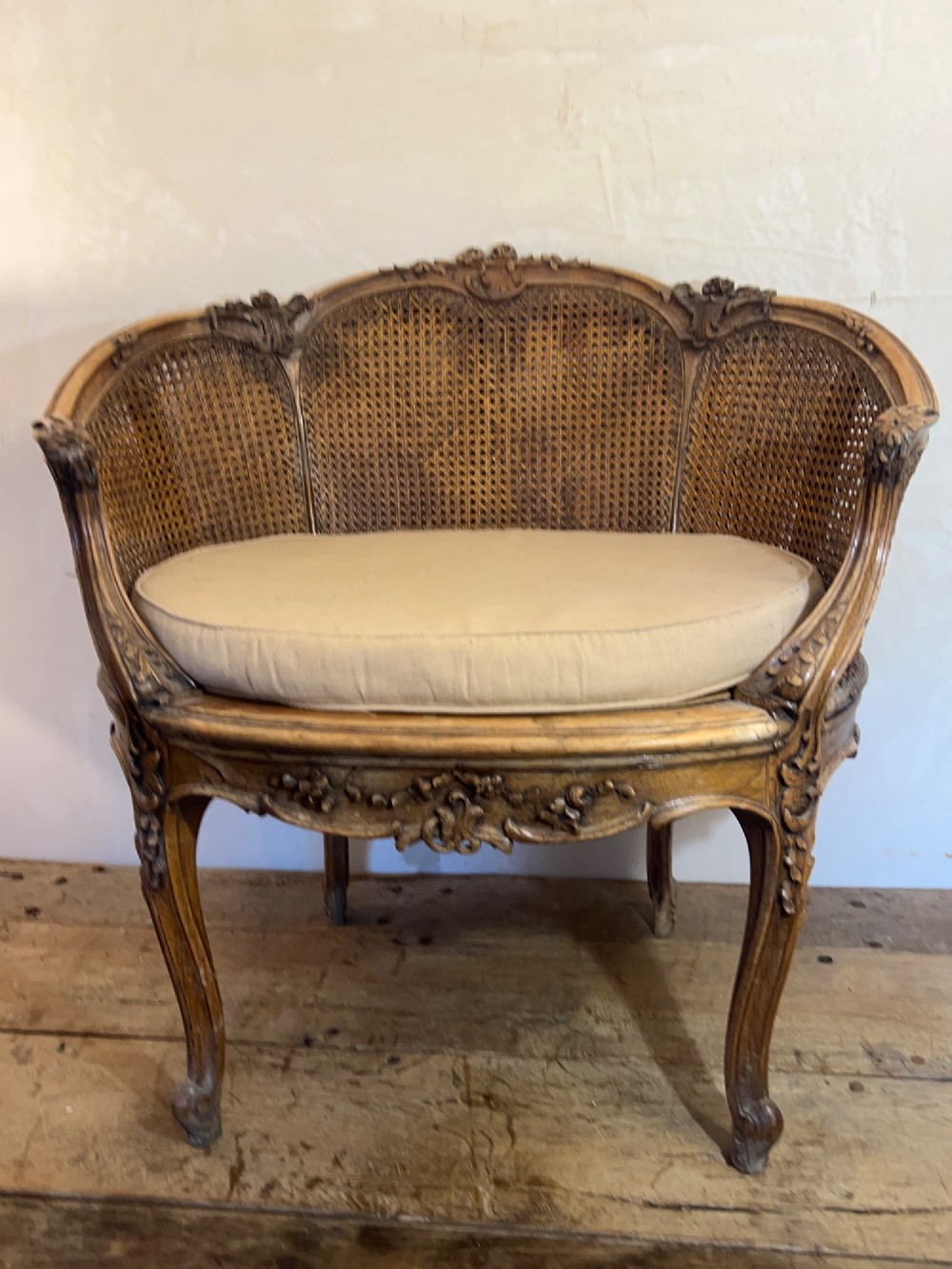 A 19th-century French Louis XV style Corbeille armchair featuring a curved hand-woven cane backrest, carved wooden frame with cabriole legs, and a beige upholstered seat cushion. View XV