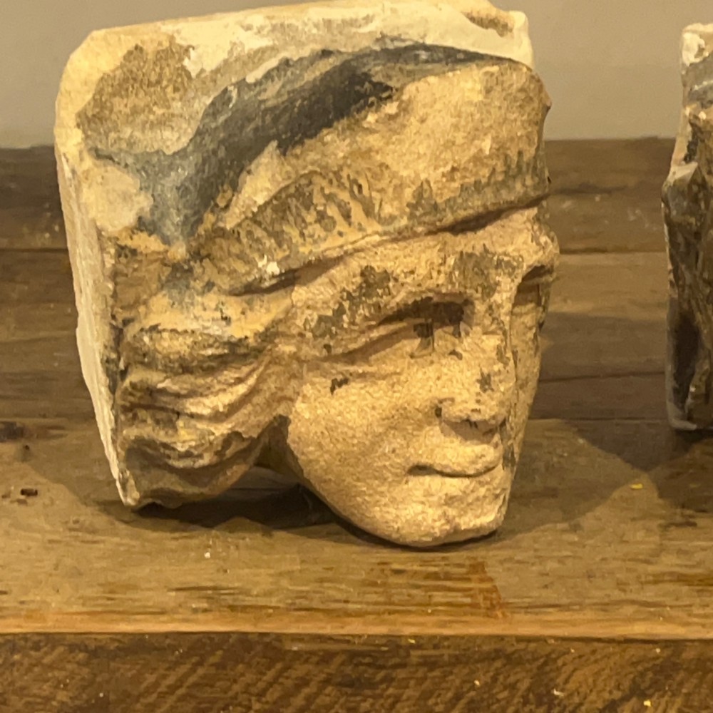 A pair of 15th-century carved limestone corbels depicting human heads with weathered facial features and period headwear. View X