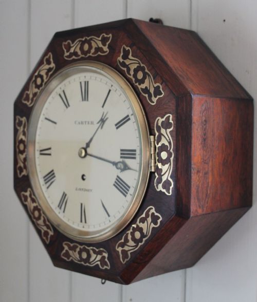 fig 1. A Regency octagonal wall clock featuring a dark wood surround decorated with brass floral inlay and an off-white dial with Roman numerals.