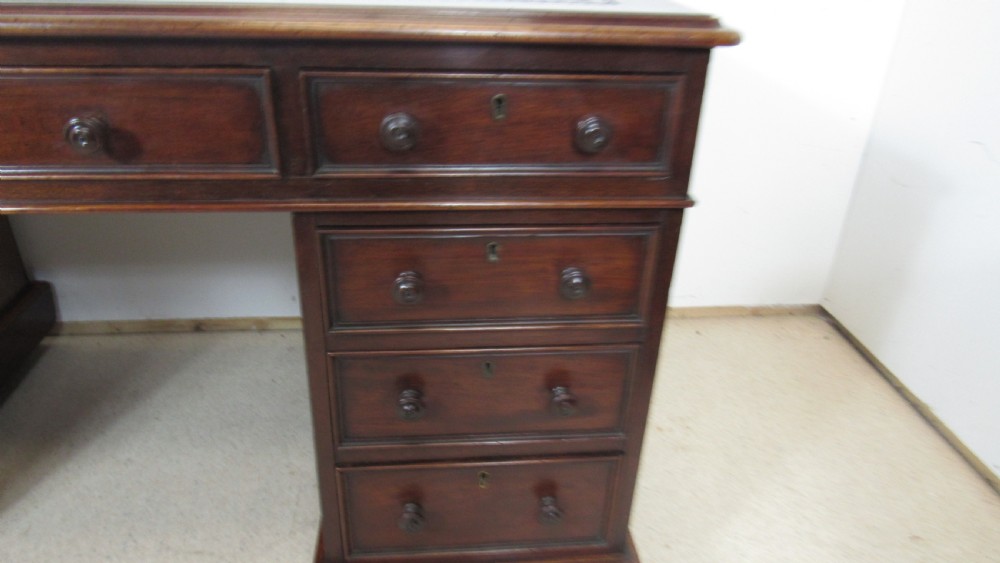 A dark mahogany pedestal desk featuring an inset leather writing surface, multiple drawers fitted with turned wooden knobs, and a central kneehole. View VI