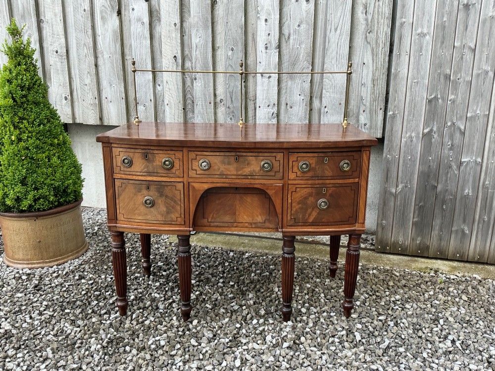 A Regency mahogany bow-front sideboard featuring reeded legs, inlaid drawer fronts with brass ring handles, and a slim brass curtain rail on the top surface.