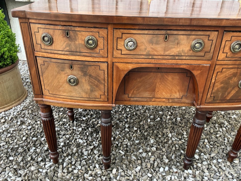 A Regency mahogany bow-front sideboard featuring reeded legs, inlaid drawer fronts with brass ring handles, and a slim brass curtain rail on the top surface. View II
