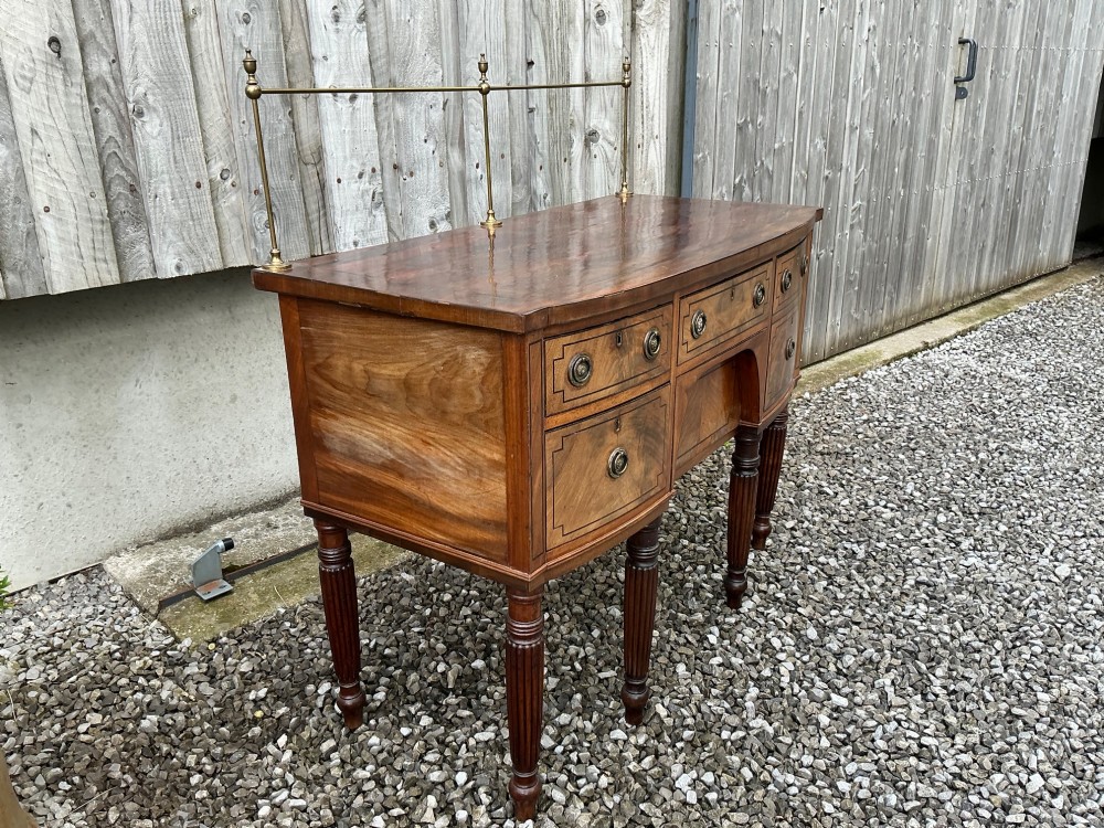 A Regency mahogany bow-front sideboard featuring reeded legs, inlaid drawer fronts with brass ring handles, and a slim brass curtain rail on the top surface. View IV