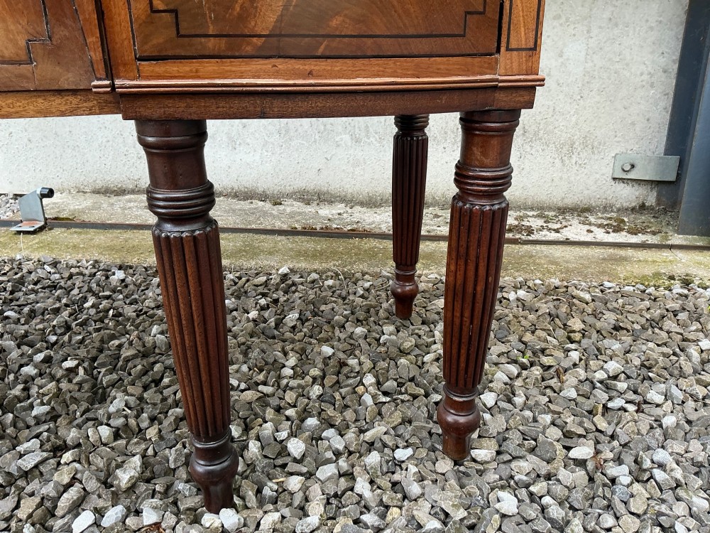 A Regency mahogany bow-front sideboard featuring reeded legs, inlaid drawer fronts with brass ring handles, and a slim brass curtain rail on the top surface. View V