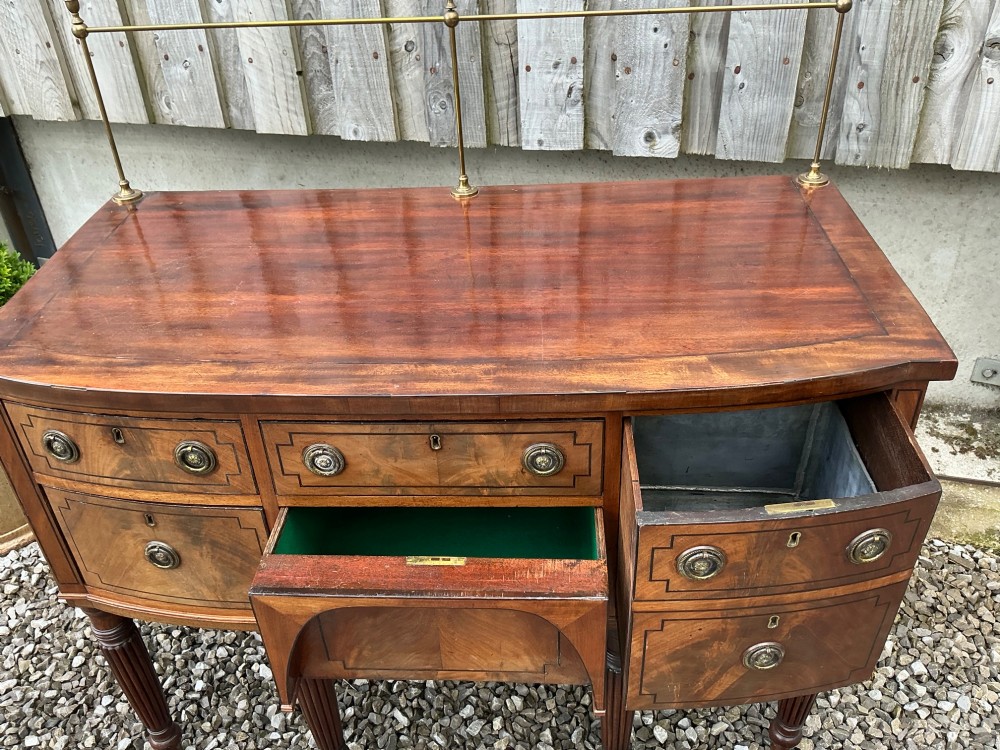 A Regency mahogany bow-front sideboard featuring reeded legs, inlaid drawer fronts with brass ring handles, and a slim brass curtain rail on the top surface. View VI