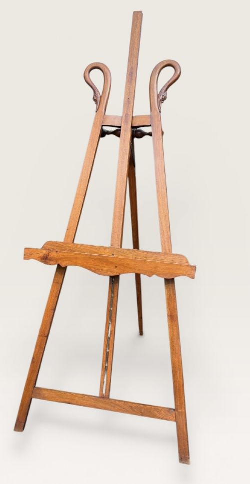 A tall warm brown wooden floor-standing tripod easel featuring carved swan head finials at the top arch and a wide horizontal support shelf. View VI