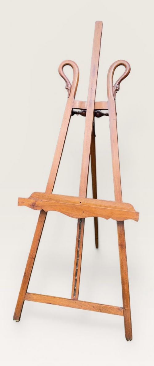 A tall warm brown wooden floor-standing tripod easel featuring carved swan head finials at the top arch and a wide horizontal support shelf. View VII