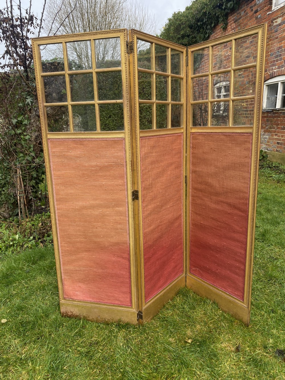 A three-panel giltwood folding room divider featuring glazed upper glass panes and pinkish terracotta fabric lower panels.