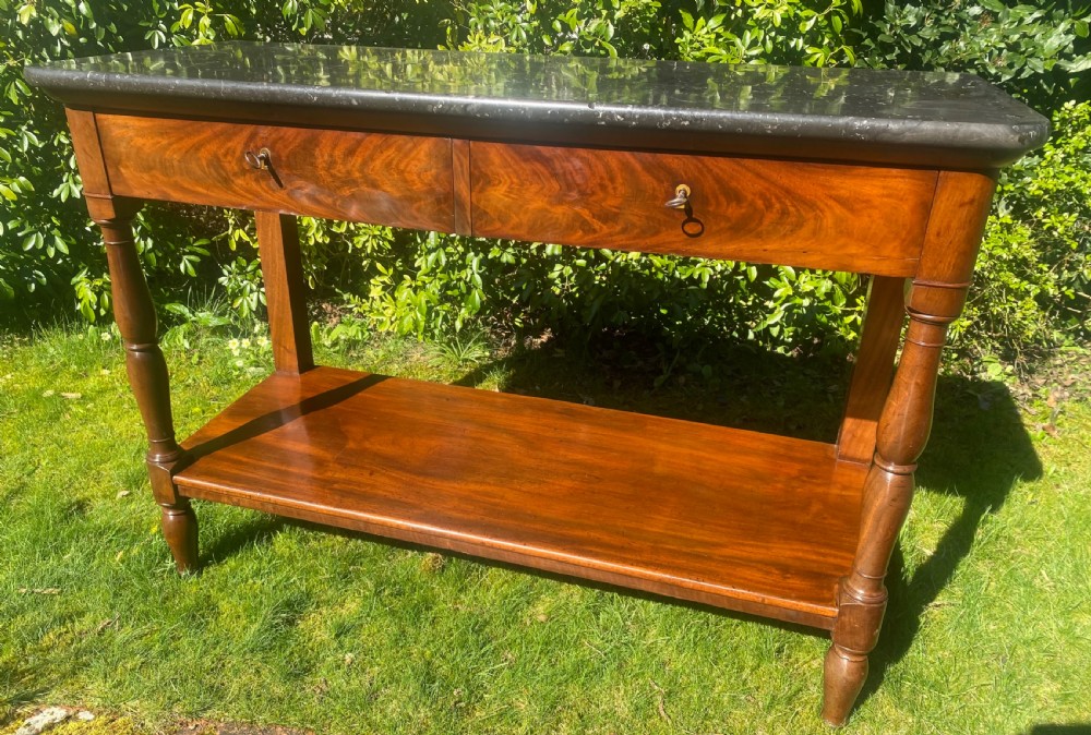 A flame mahogany console table featuring a dark grey marble slab top, two frieze drawers with brass keyholes, turned front supports, and a low undertier shelf.