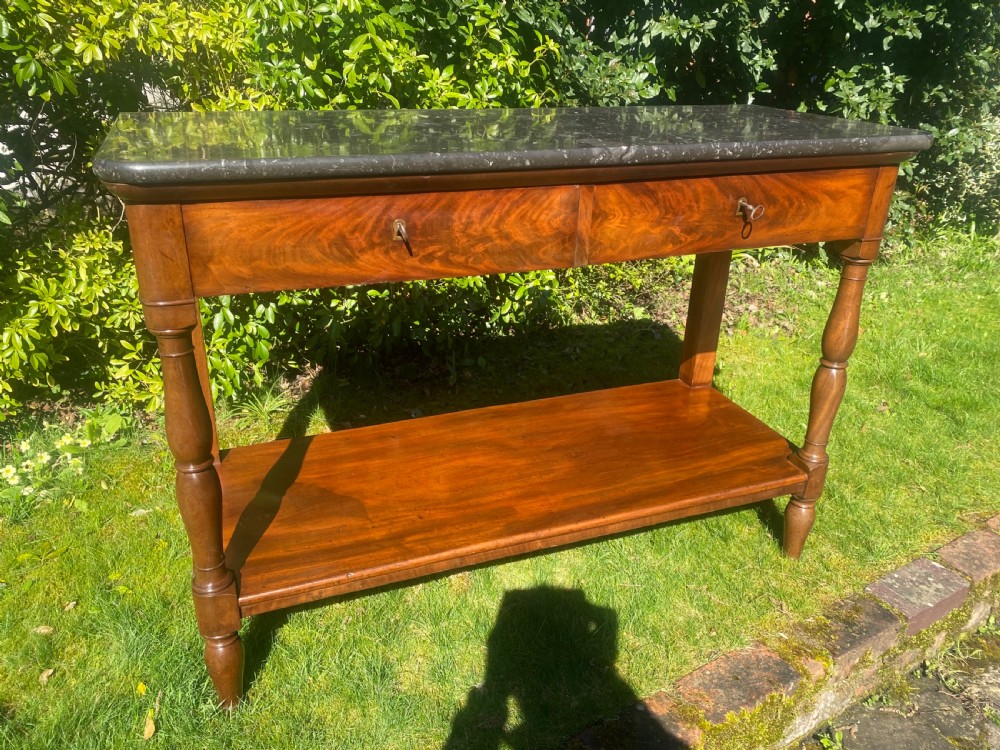 A flame mahogany console table featuring a dark grey marble slab top, two frieze drawers with brass keyholes, turned front supports, and a low undertier shelf. View VI