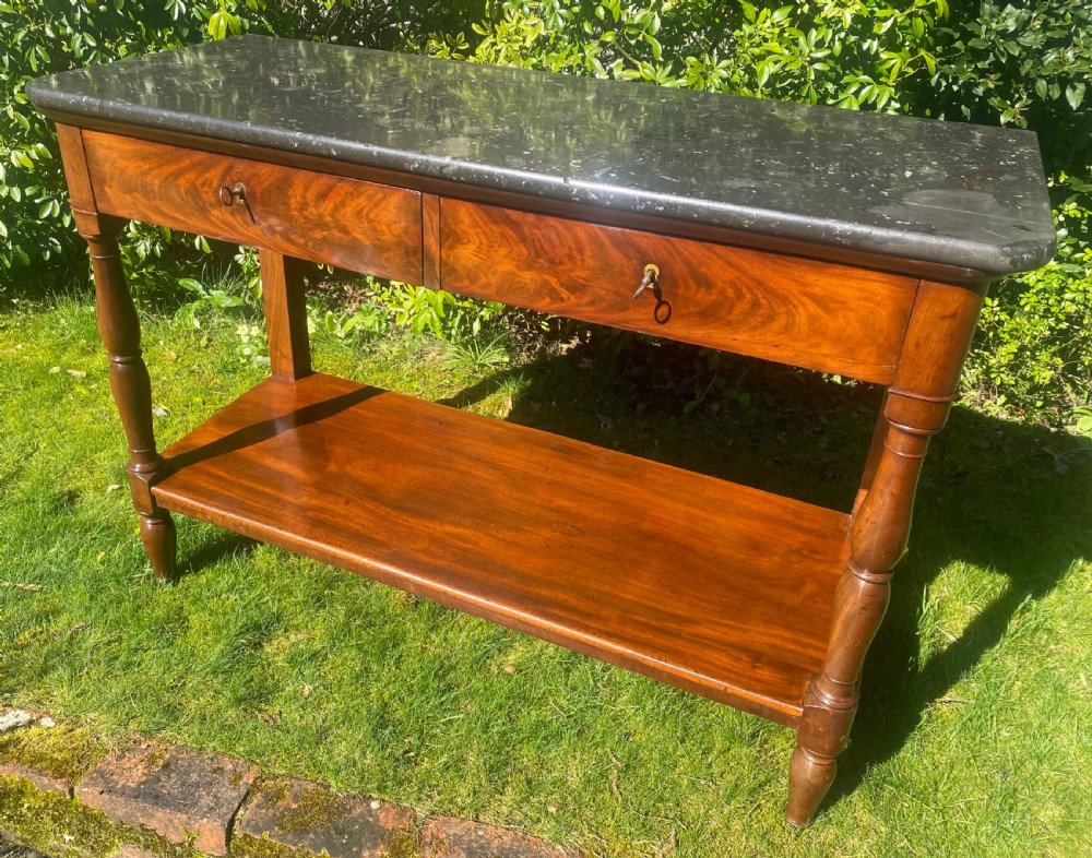 A flame mahogany console table featuring a dark grey marble slab top, two frieze drawers with brass keyholes, turned front supports, and a low undertier shelf. View VII
