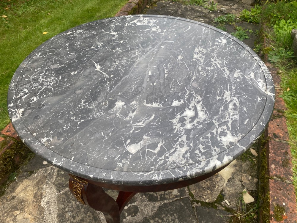 A French Restauration period mahogany gueridon table featuring a circular grey marble top, three curved legs with gilt bronze mounts, and a lower triangular stretcher. View VII