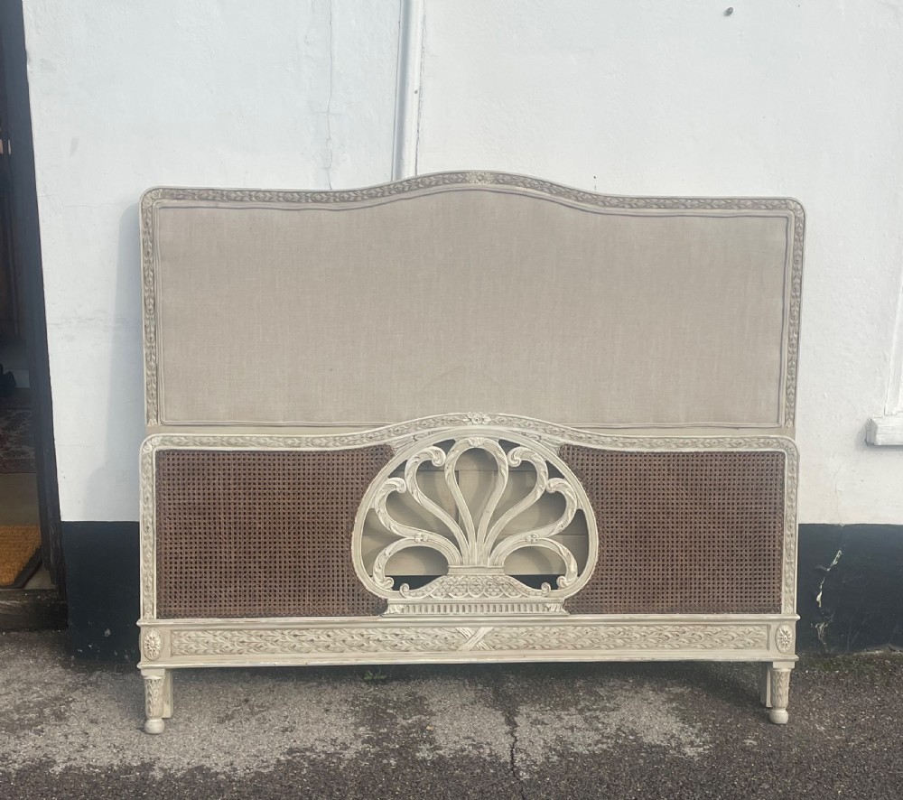 A carved cream painted king size bed headboard with a beige upholstered upper section and woven cane lower section featuring a central carved fan motif.