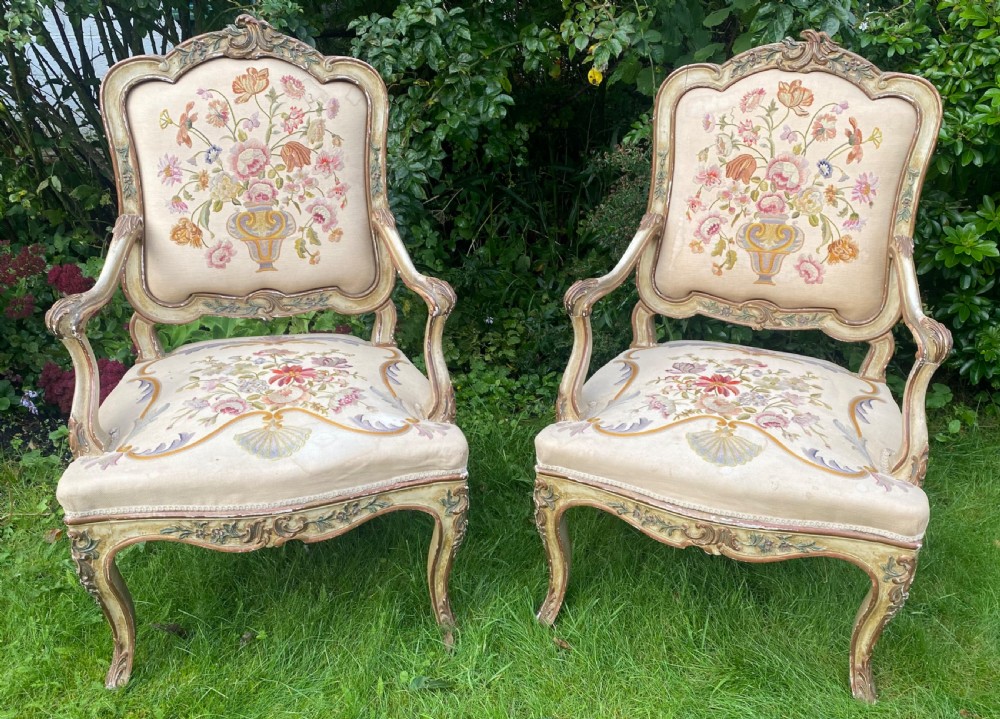 A pair of Italian Rococo style armchairs featuring cream-painted carved wooden frames and floral needlework upholstery depicting urns filled with flowers.