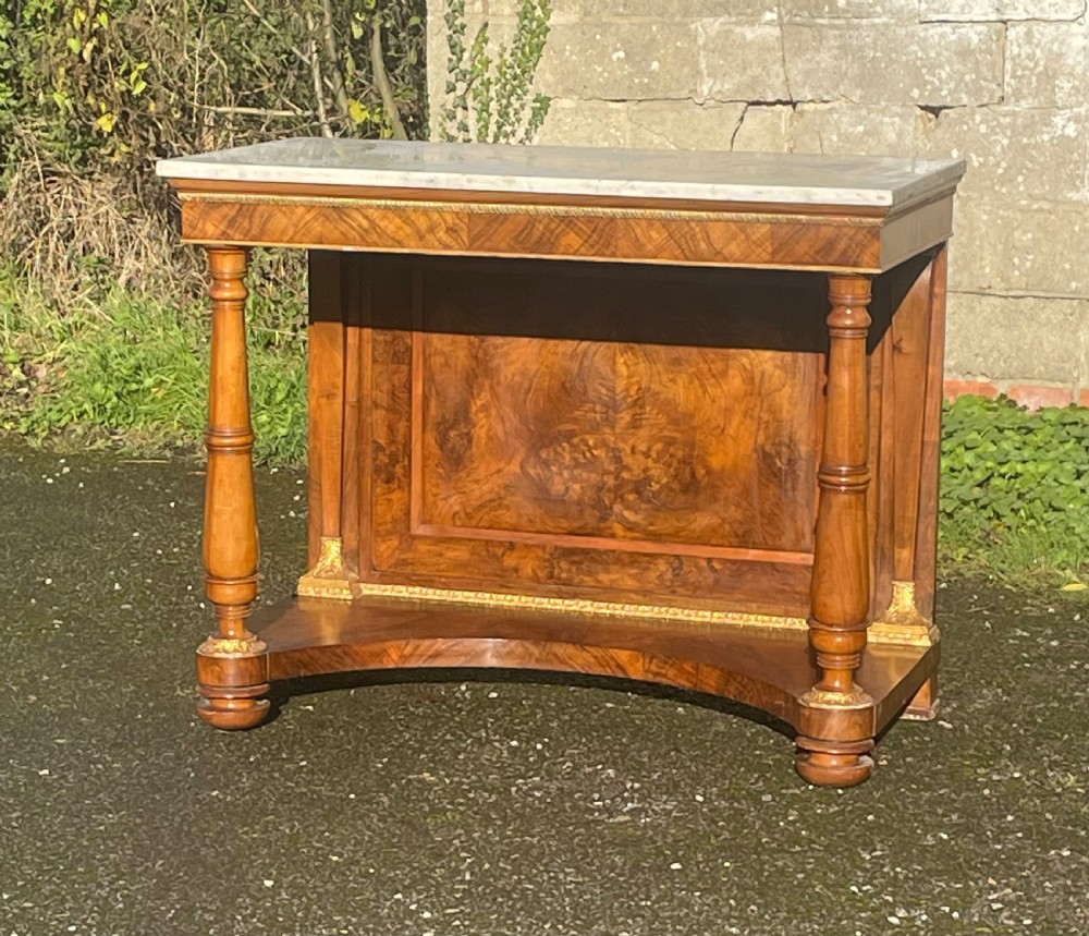 A 19th-century French Empire style walnut console table featuring a grey marble top, turned column supports with gilt metal mounts, and a shaped lower shelf.
