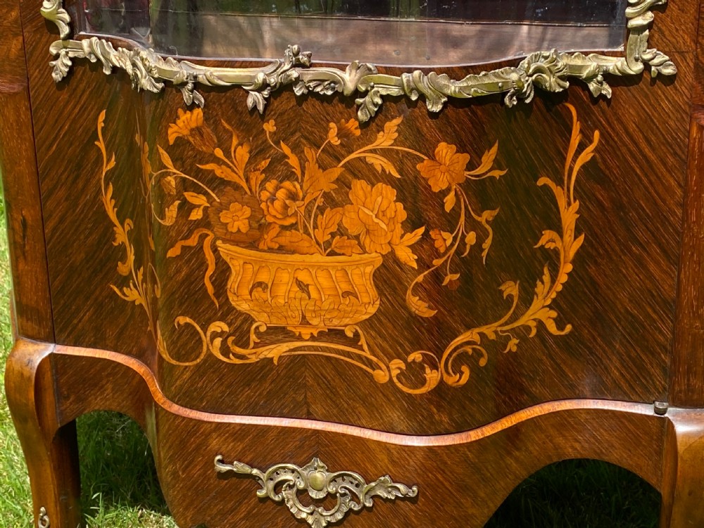 A French Rococo style serpentine glazed vitrine cabinet featuring dark wood veneer, intricate floral marquetry inlay on the lower panel, and decorative gilt bronze ormolu mounts. View V