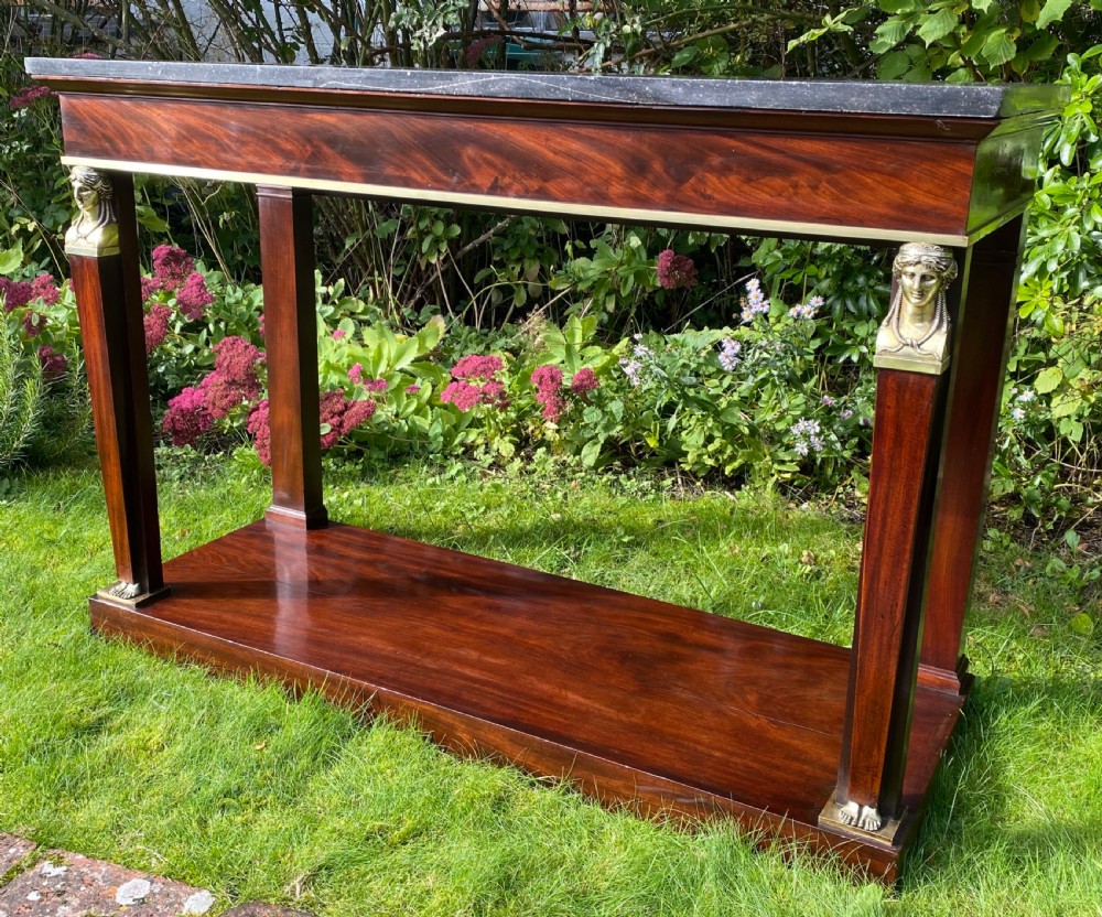 A French Empire console table featuring flame mahogany veneer, a dark marble top, gilt bronze caryatid mounts, pilaster legs, and a lower plinth shelf. View II
