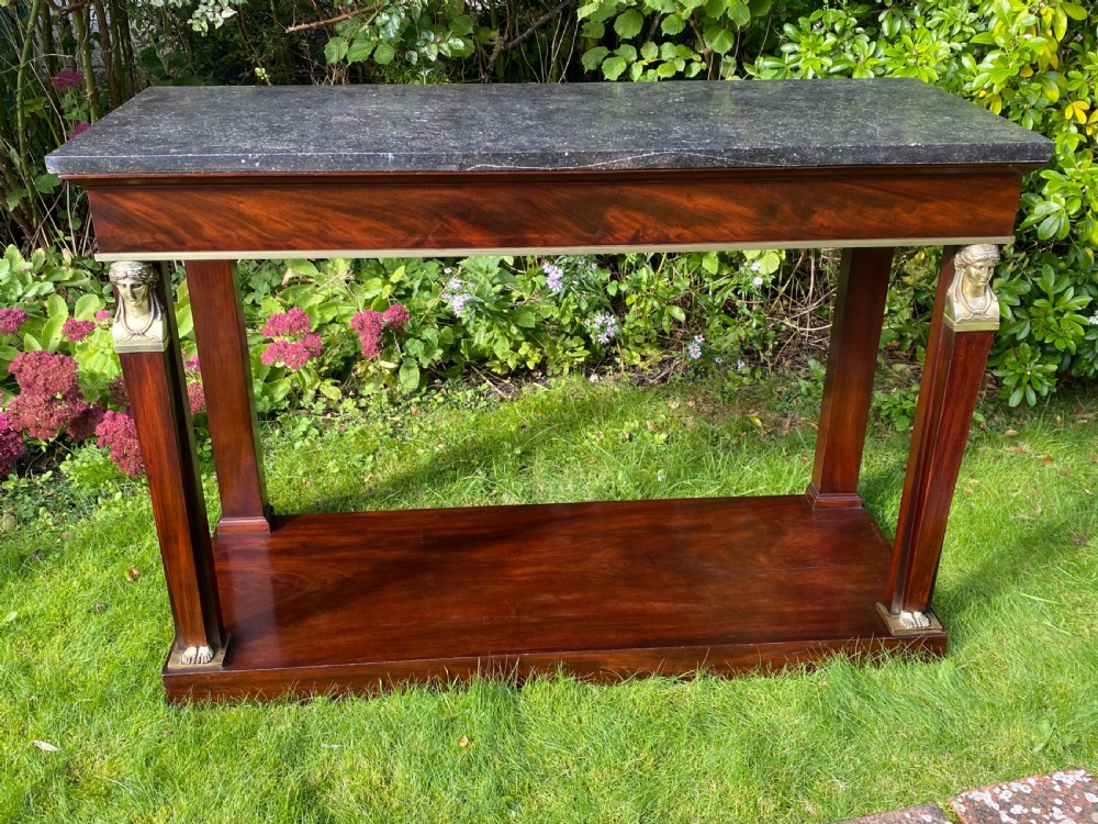 A French Empire console table featuring flame mahogany veneer, a dark marble top, gilt bronze caryatid mounts, pilaster legs, and a lower plinth shelf. View V