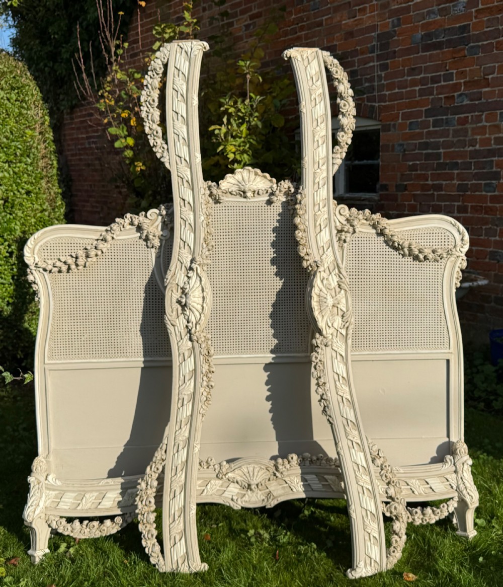 A cream painted kingsize bed frame components featuring carved floral garlands, shell cresting, and woven rattan cane panels.