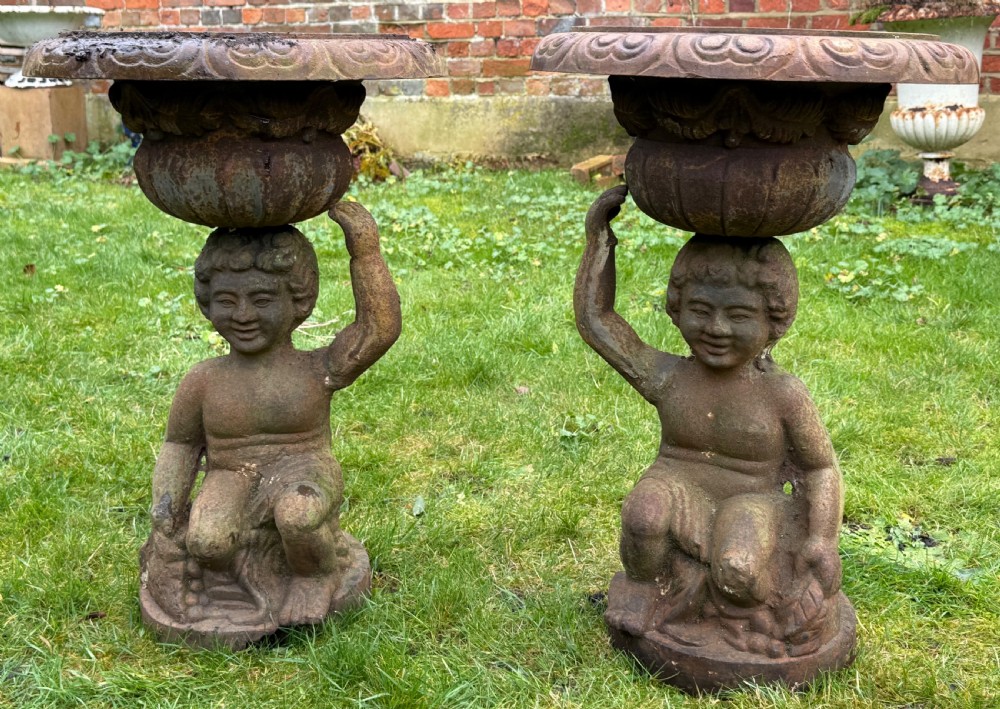 A pair of weathered rust-brown cast iron garden planters featuring cherub figures holding wide gadrooned urn bowls.