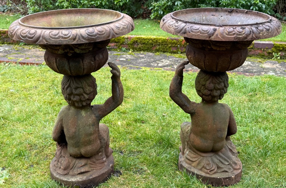 A pair of weathered rust-brown cast iron garden planters featuring cherub figures holding wide gadrooned urn bowls. View III