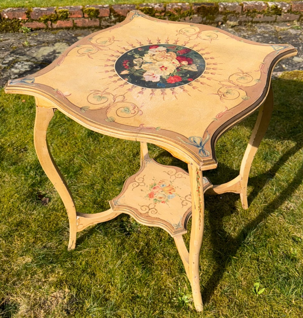 A cream-painted two-tier occasional table featuring cabriole legs, a shaped serpentine top with a floral central medallion, and decorative scrollwork borders.