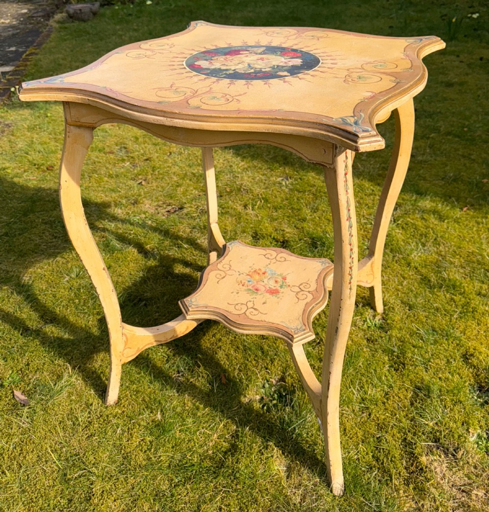 A cream-painted two-tier occasional table featuring cabriole legs, a shaped serpentine top with a floral central medallion, and decorative scrollwork borders. View VI