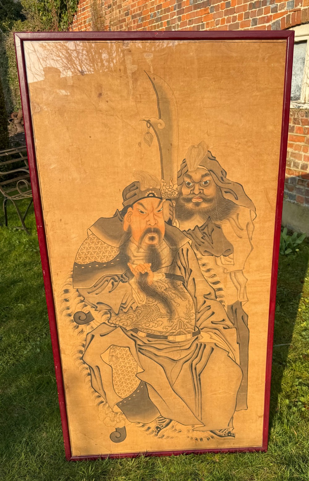 A large framed Chinese ink and watercolour painting depicting a seated bearded general accompanied by an attendant holding a crescent blade against an ochre background.