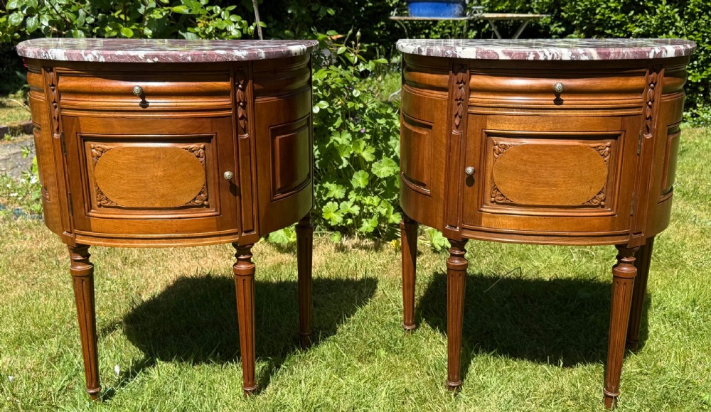 Pair of semi-circular walnut bedside cabinets with carved door panels, fluted legs, and variegated marble tops.
