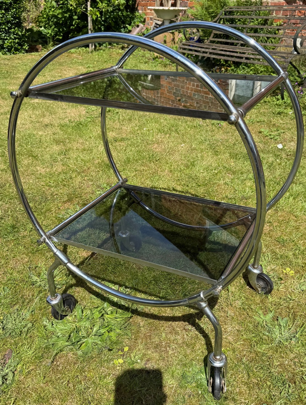 An Art Deco circular chrome frame drinks trolley with two rectangular smoked glass shelves set on castor wheels.