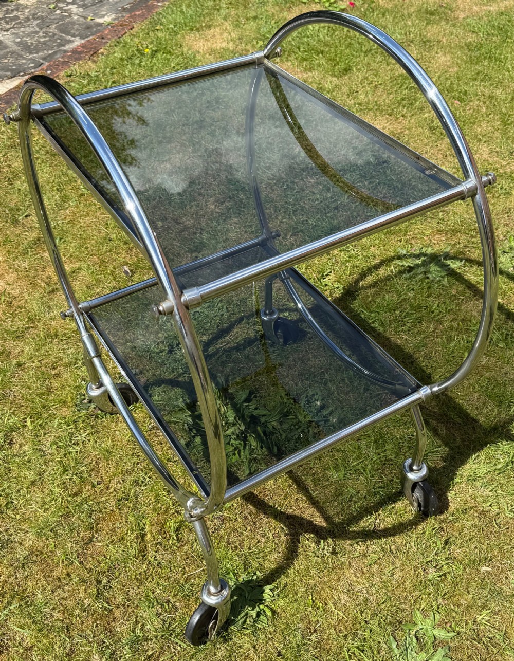 An Art Deco circular chrome frame drinks trolley with two rectangular smoked glass shelves set on castor wheels. View V