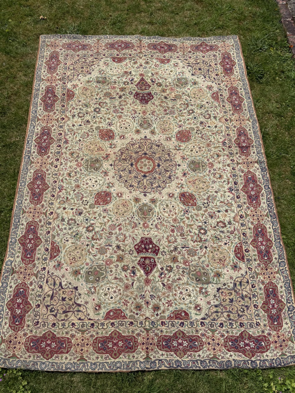 A rectangular hand-embroidered crewelwork carpet featuring a central medallion and intricate floral motifs in burgundy, green, and navy blue on a cream ground.