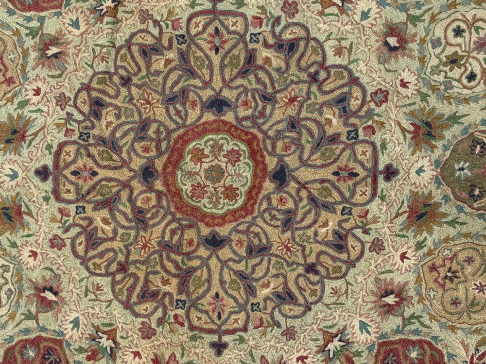 A rectangular hand-embroidered crewelwork carpet featuring a central medallion and intricate floral motifs in burgundy, green, and navy blue on a cream ground. View II