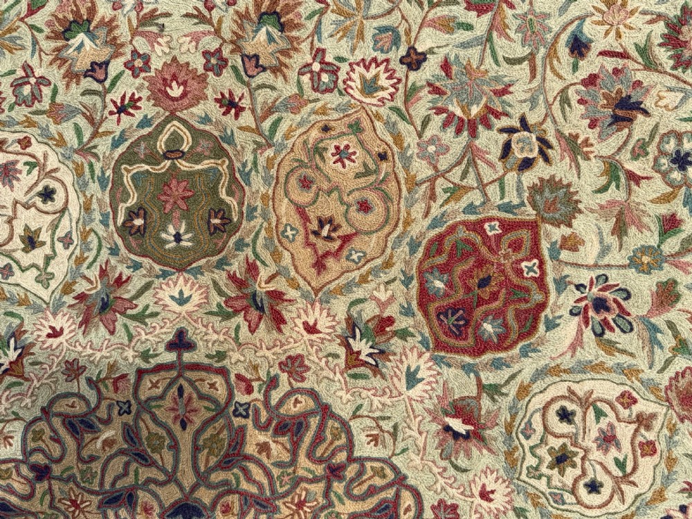 A rectangular hand-embroidered crewelwork carpet featuring a central medallion and intricate floral motifs in burgundy, green, and navy blue on a cream ground. View III