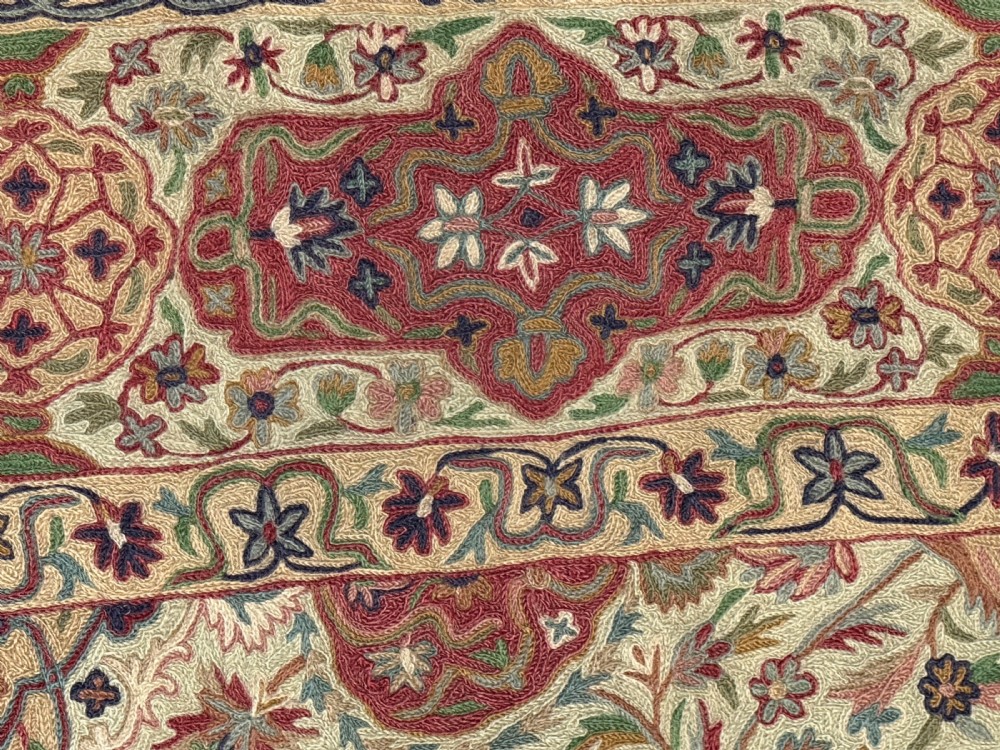 A rectangular hand-embroidered crewelwork carpet featuring a central medallion and intricate floral motifs in burgundy, green, and navy blue on a cream ground. View V