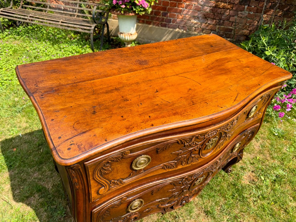 An 18th-century French Provencal walnut commode featuring a serpentine front, intricate ribbon and foliage carvings, two drawers with brass ring handles, and cabriole legs. View VI