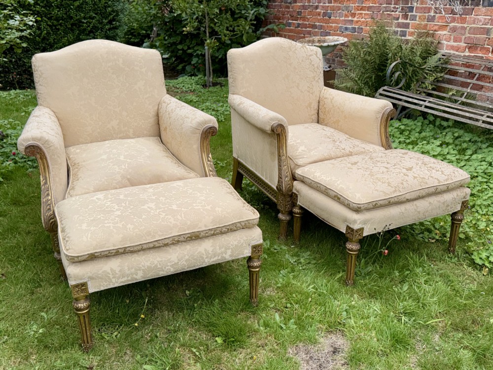 A pair of giltwood armchairs upholstered in cream damask pattern fabric with matching footstools.
