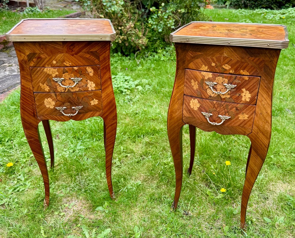 A pair of French Louis XV style marquetry bedside tables with floral wood inlays, brass rimmed tops, twin drawers, and slender cabriole legs.