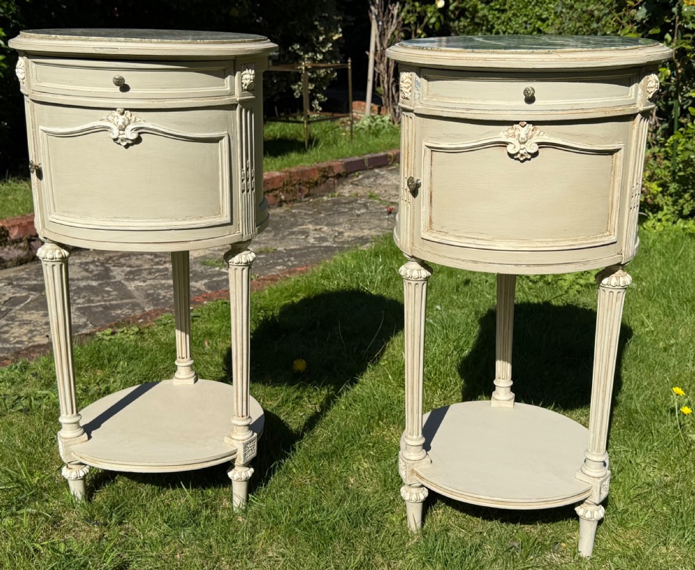 A pair of oval off-white painted Louis XVI style bedside tables featuring marble tops, single drawers, cupboard doors with carved details, and fluted legs with a lower shelf.