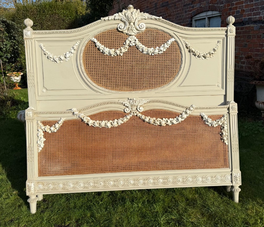 A cream-painted king-size bedstead featuring carved floral garlands, fluted corner posts, and woven natural cane panels on the headboard and footboard.
