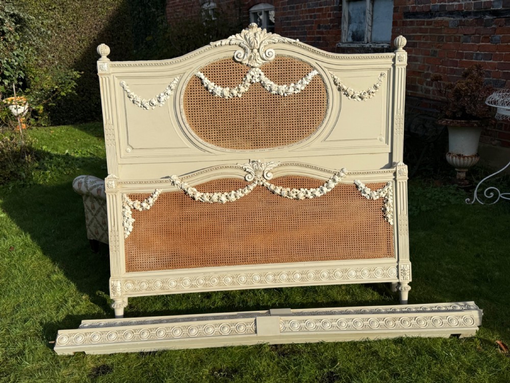 A cream-painted king-size bedstead featuring carved floral garlands, fluted corner posts, and woven natural cane panels on the headboard and footboard. View II