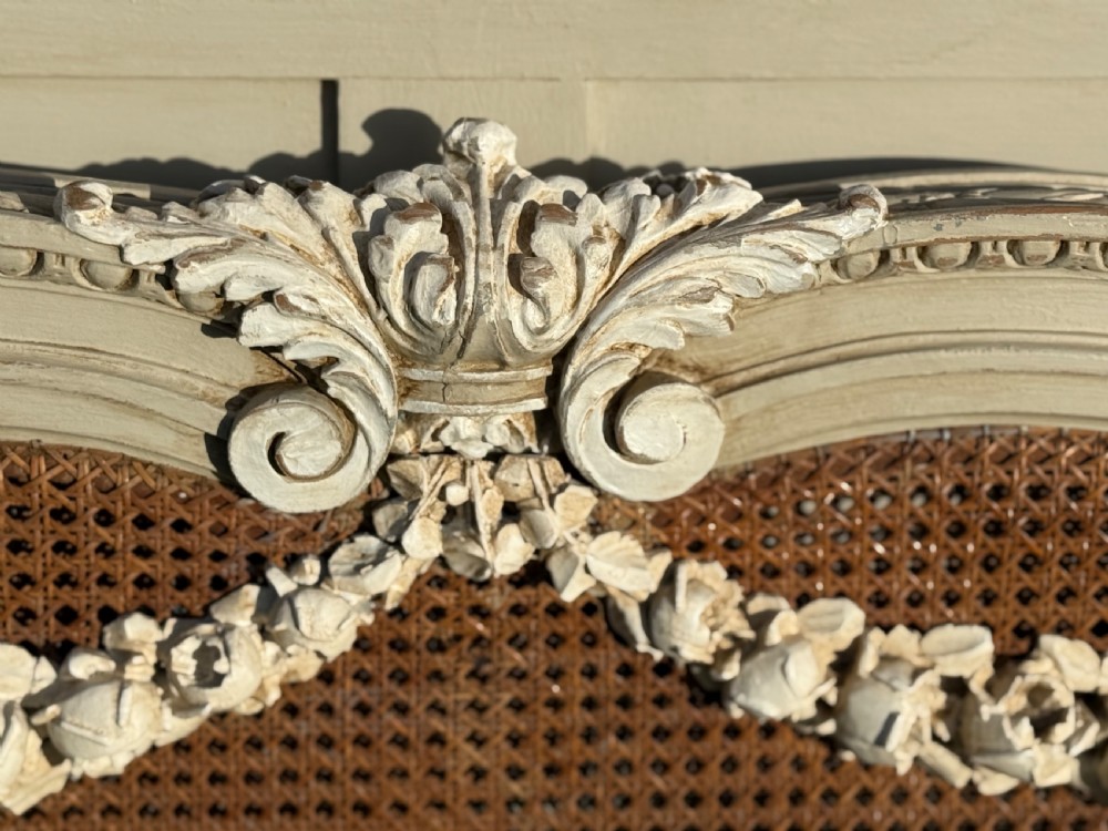 A cream-painted king-size bedstead featuring carved floral garlands, fluted corner posts, and woven natural cane panels on the headboard and footboard. View III