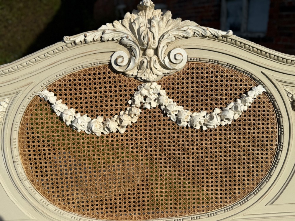 A cream-painted king-size bedstead featuring carved floral garlands, fluted corner posts, and woven natural cane panels on the headboard and footboard. View IV