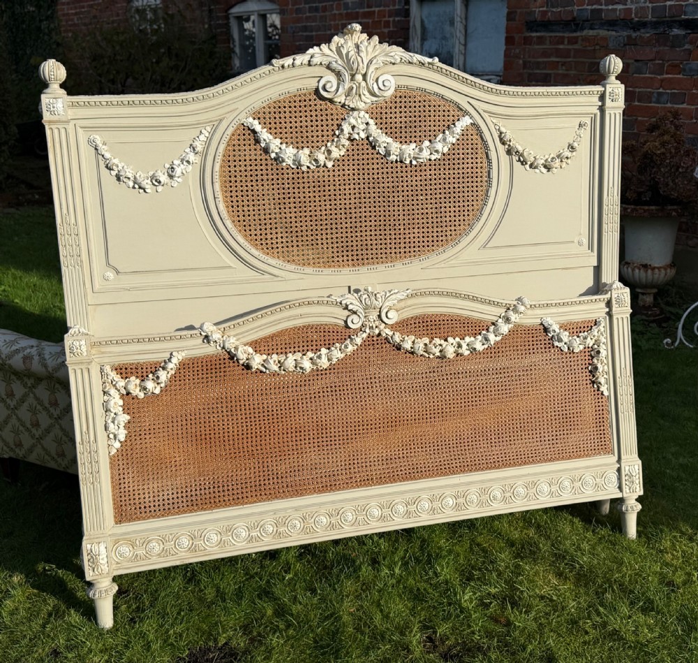 A cream-painted king-size bedstead featuring carved floral garlands, fluted corner posts, and woven natural cane panels on the headboard and footboard. View V