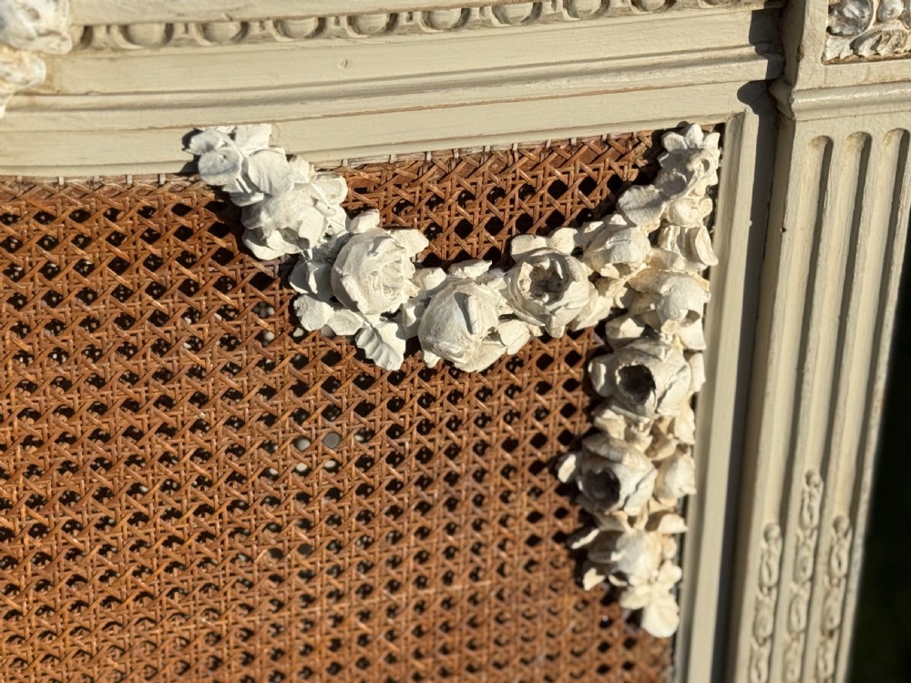 A cream-painted king-size bedstead featuring carved floral garlands, fluted corner posts, and woven natural cane panels on the headboard and footboard. View VI
