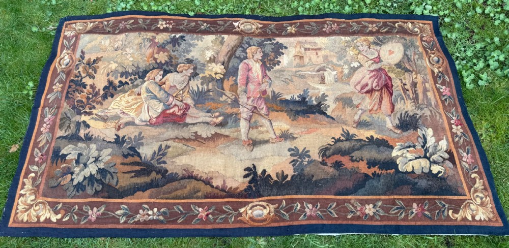 A 19th-century woven wall tapestry depicting a pastoral landscape scene with figures in romantic attire within a detailed floral border. View II