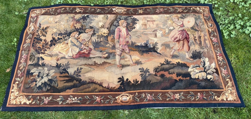 A 19th-century woven wall tapestry depicting a pastoral landscape scene with figures in romantic attire within a detailed floral border.