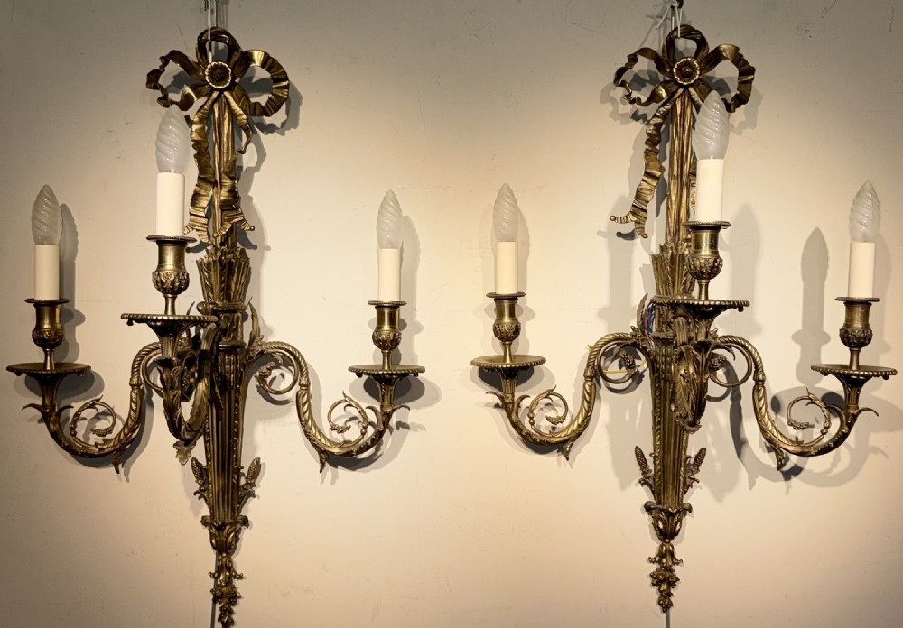 A pair of cast brass three-light wall sconces featuring ribbon bow tops and acanthus leaf scrolled arms.