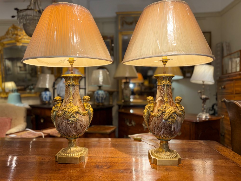 A pair of large table lamps featuring brown veined marble urn-shaped bases adorned with ornate gilt bronze mounts and fitted with pleated fabric shades.